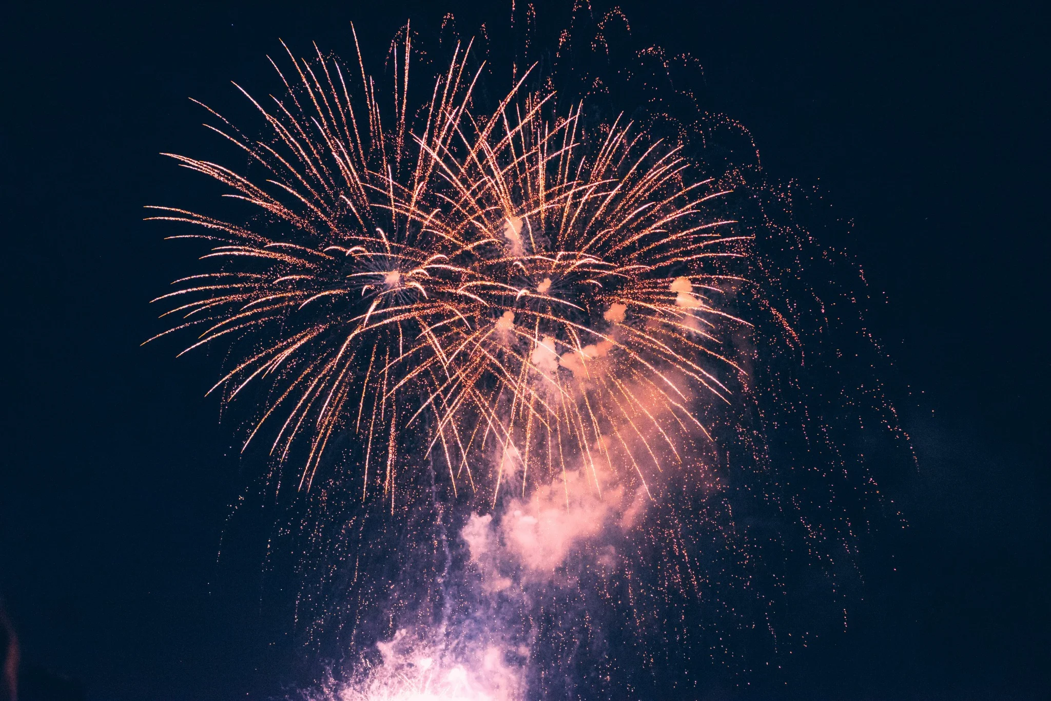 Fuegos artificiales dorados iluminando el cielo nocturno durante una celebración, con destellos brillantes y una nube de humo en la parte inferior de la explosión.